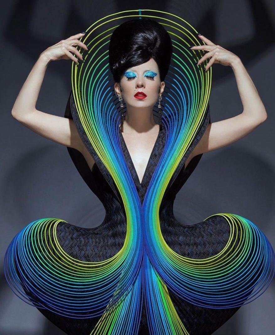 A woman with dark hair poses in a sculptural black gown with yellow, blue, and green accents against a gray background in a studio shot.