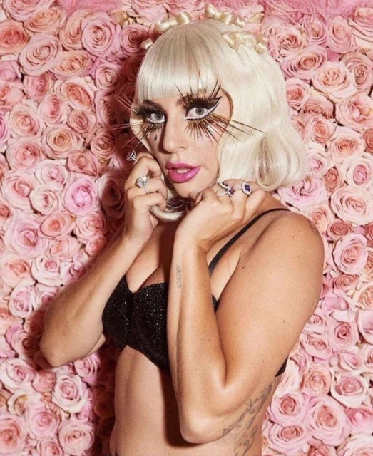Woman with platinum blonde bob and large eyelashes poses against a backdrop of pink roses, wearing a dark bra top and rings.