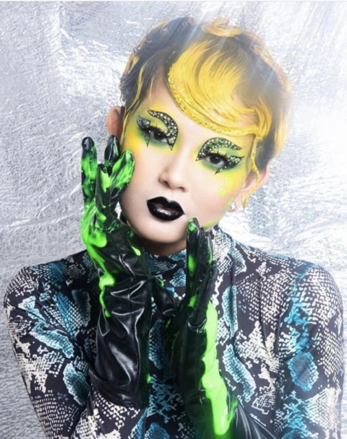 Close-up of a model with stylized makeup and green gloves against a silver background, featuring a snakeskin-print top.