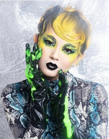 Close-up of a model with stylized makeup and green gloves against a silver background, featuring a snakeskin-print top.