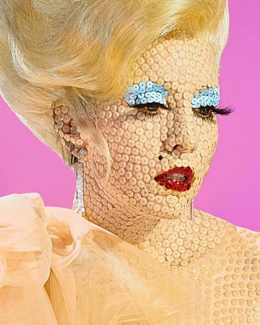 Artistic portrait of a woman with sequins covering her face, dramatic eye makeup, and a voluminous blonde wig against a pink background.