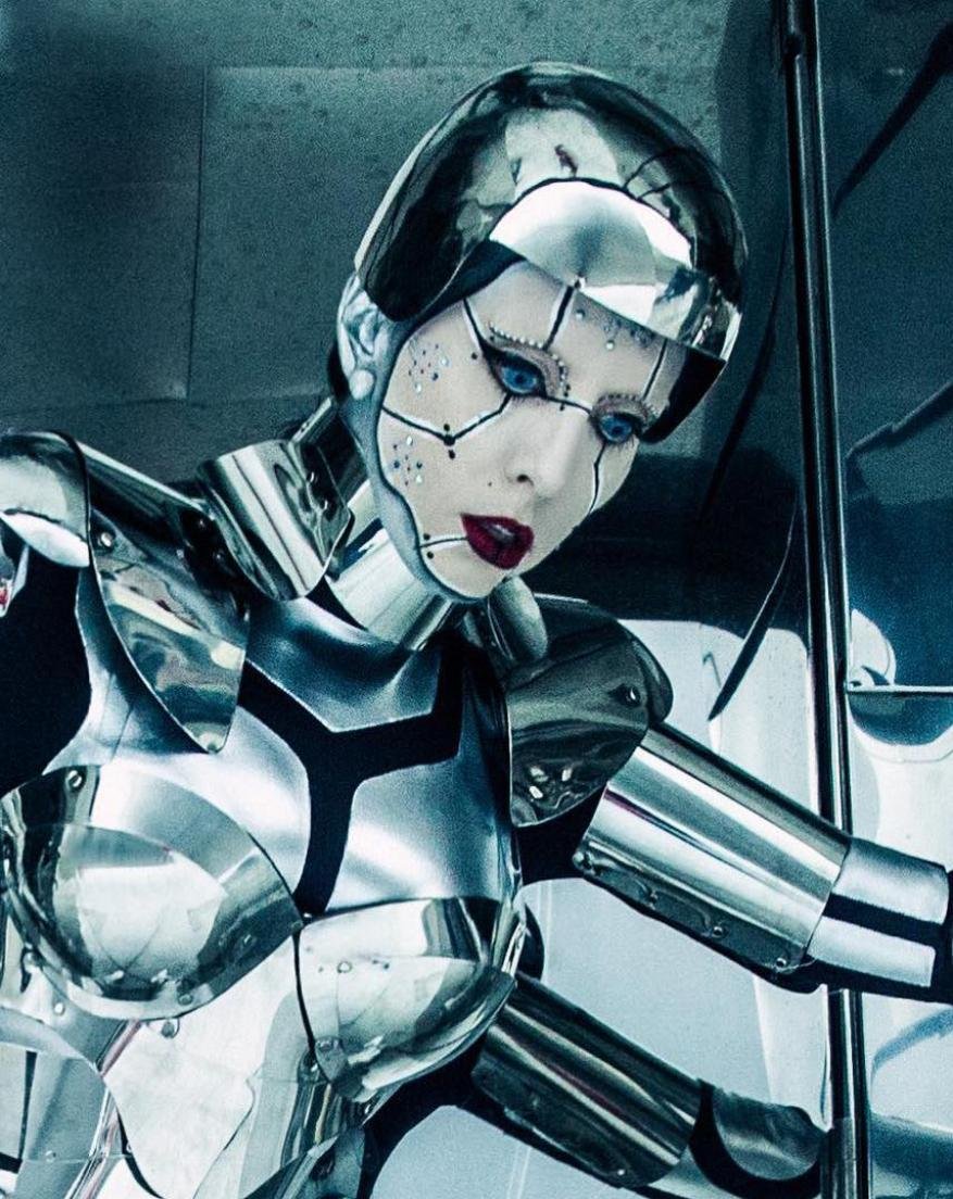 CGI portrait of a woman wearing futuristic chrome armour and helmet, with fair skin, striking blue eyes, and dark lipstick in an indoor studio setting.