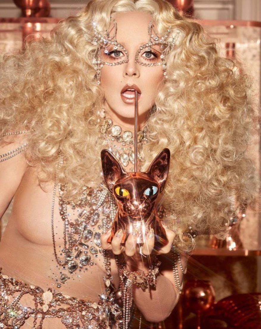 Glamorous woman with curly blonde hair and jeweled mask posing with a copper cat statue, wearing sparkling jewelry and sheer clothing.
