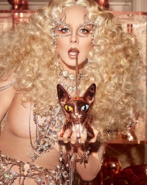 Glamorous woman with curly blonde hair and jeweled mask posing with a copper cat statue, wearing sparkling jewelry and sheer clothing.