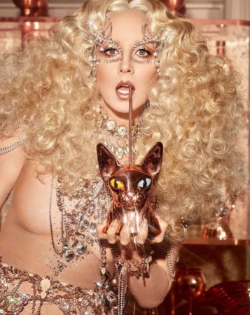 Glamorous woman with curly blonde hair and jeweled mask posing with a copper cat statue, wearing sparkling jewelry and sheer clothing.