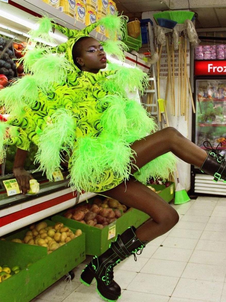 A person in a neon green feathered outfit poses in a grocery store aisle, alongside shelves of produce and refrigerators, and tiled flooring.