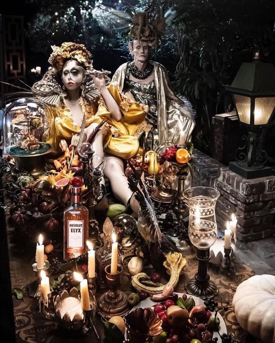Two models in ornate costumes pose behind a lavishly set table laden with fruit, candles, and an Absolut Elyx bottle in a dark, gothic-inspired setting.