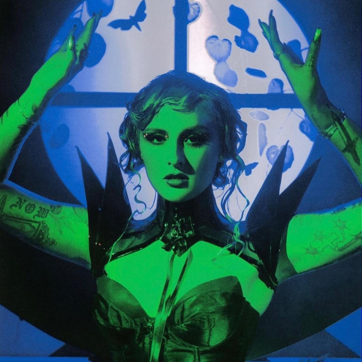A woman with green skin, dark clothing, and dramatic makeup poses in front of a window with butterfly and heart cutouts.