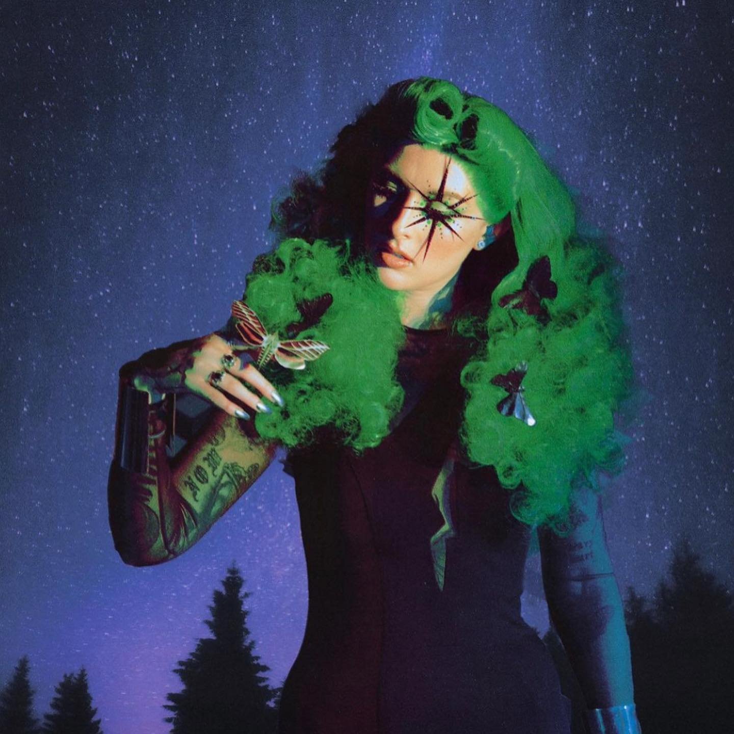 Portrait of a woman with green curly hair and face paint holding a moth in front of a night sky backdrop with evergreens.