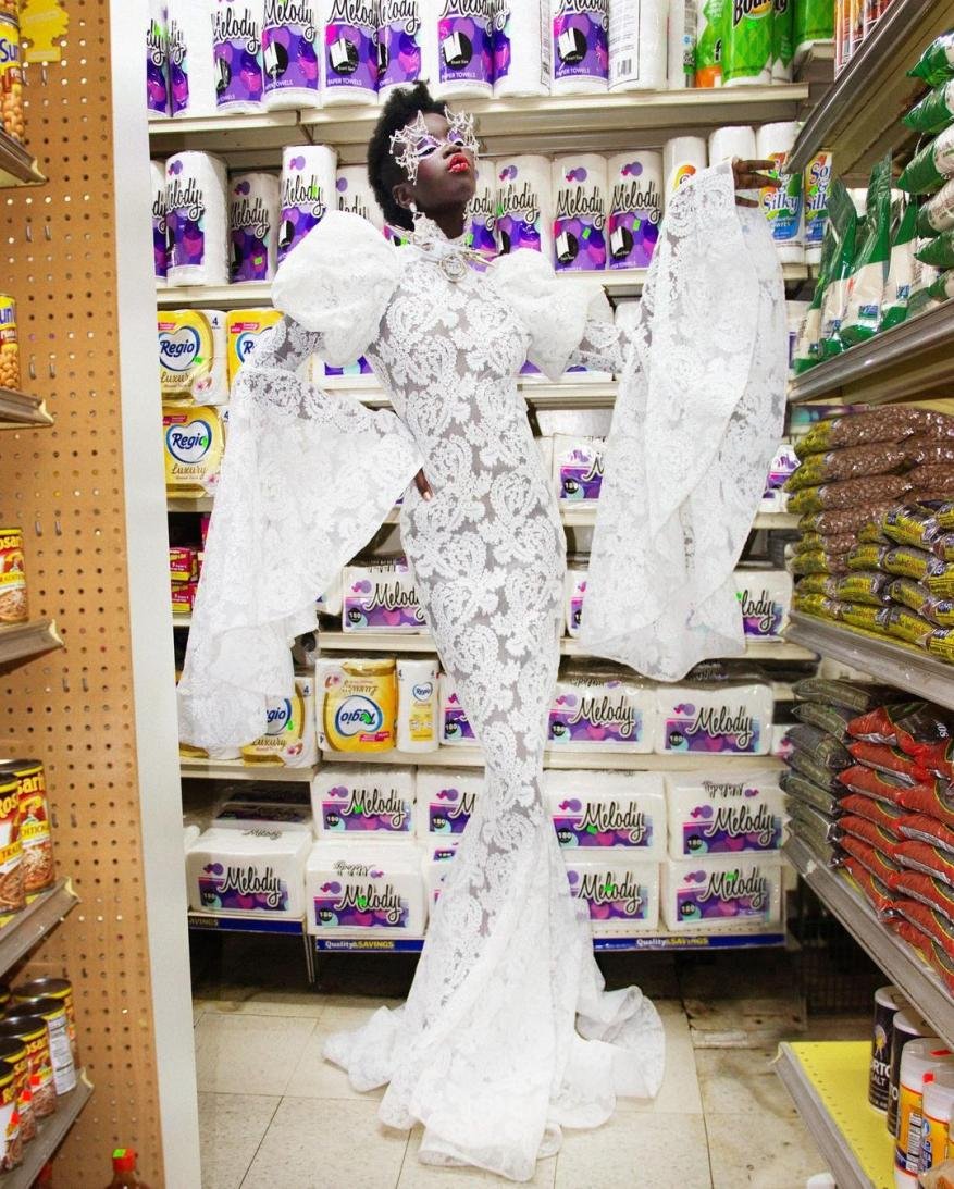 A person in a lace gown and elaborate headpiece poses in a grocery store aisle surrounded by shelves stocked with merchandise.