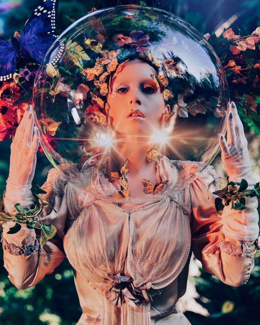 Woman with butterflies in clear bubble helmet in whimsical outdoor setting with muted colors and soft lighting. Fantasy and ethereal.
