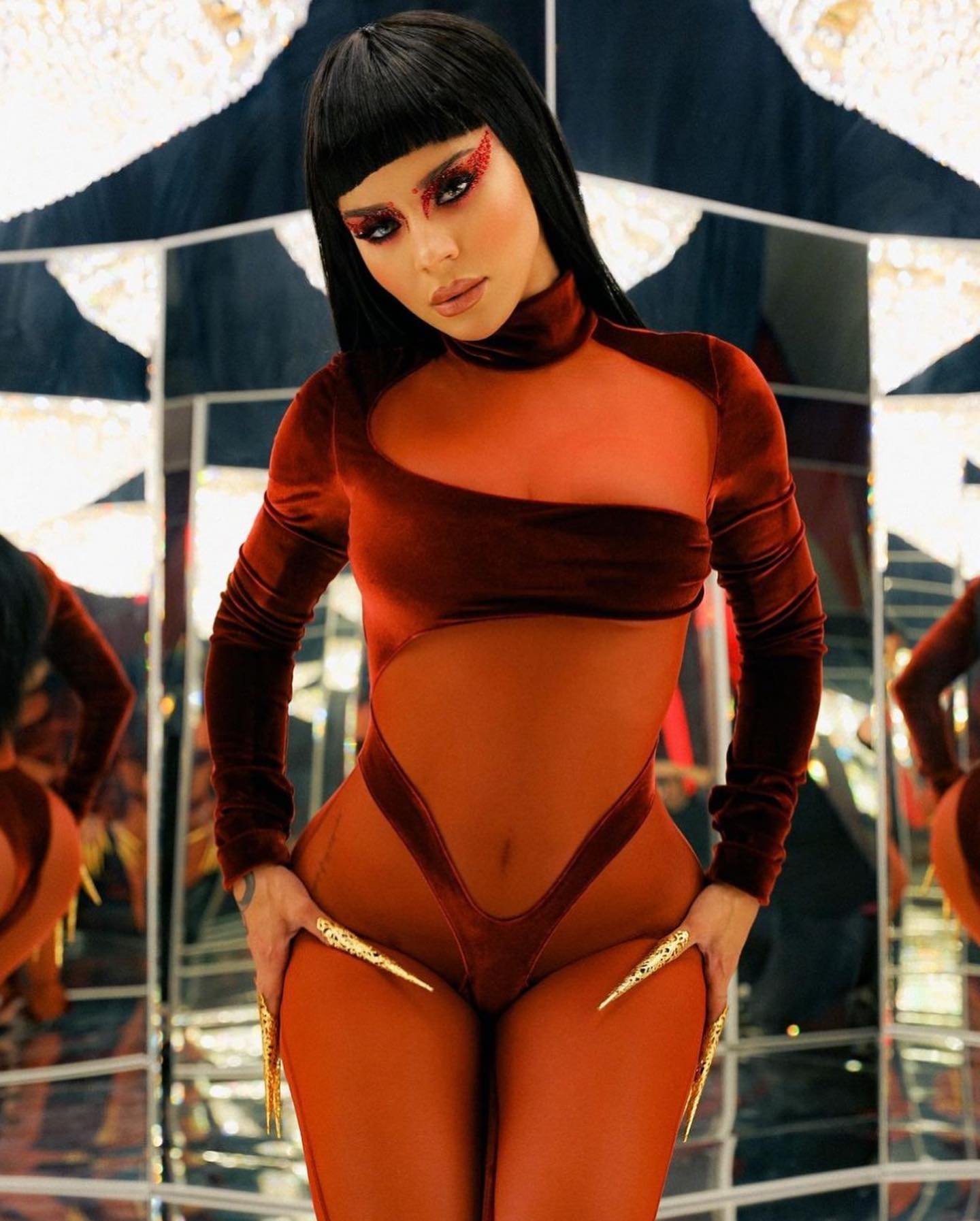 Woman in a reddish-brown velvet and sheer bodysuit with eye makeup and long nails posing in front of reflective panels in a studio.
