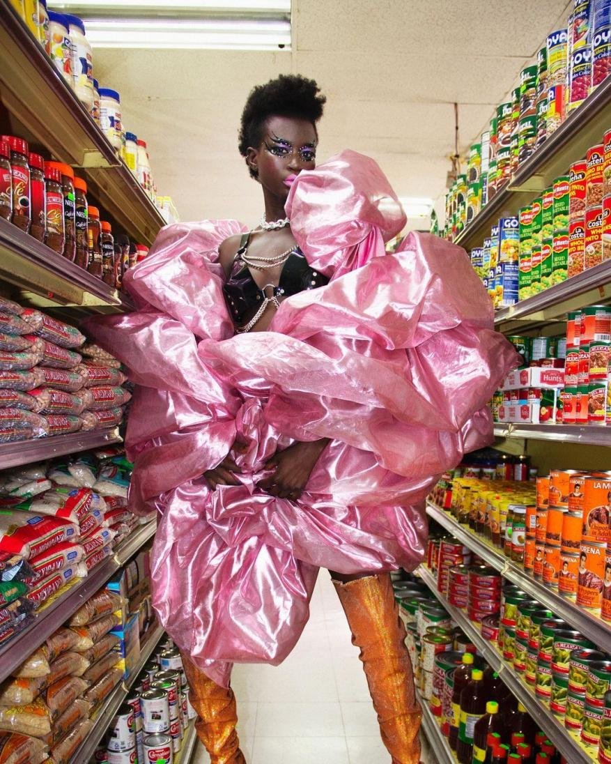 Woman in a voluminous pink dress with matching eye makeup stands in a grocery store aisle, posing among shelves of canned goods.