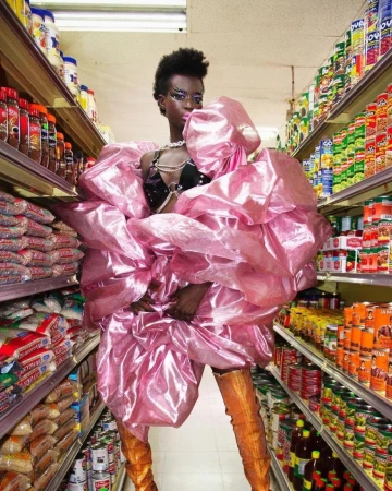 Woman in a voluminous pink dress with matching eye makeup stands in a grocery store aisle, posing among shelves of canned goods.