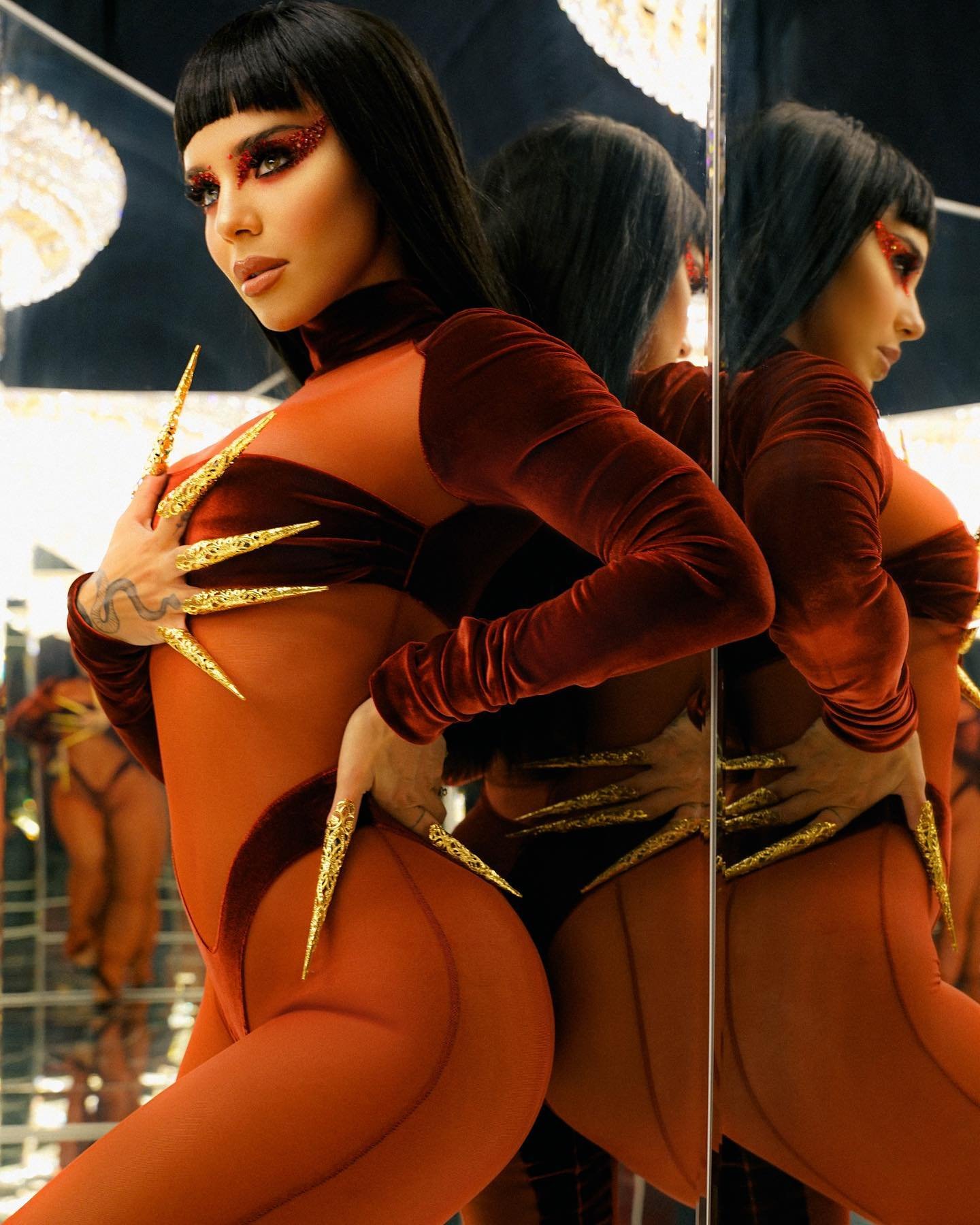 Woman in rust-colored bodysuit with long gold nails poses indoors by mirror and modern lighting, looking away in beauty shot.