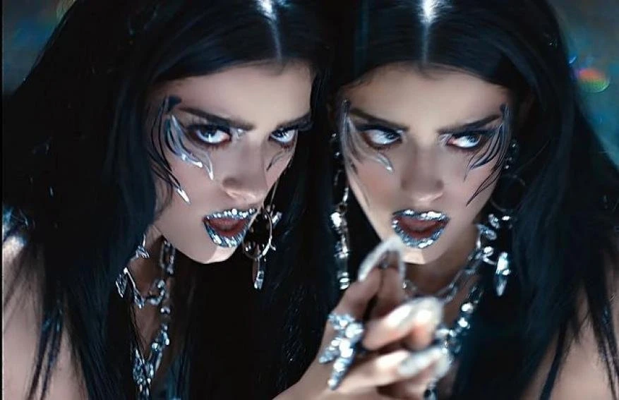 Two adult females with dramatic silver makeup and jewelry, dark hair, and intense expressions, posing in a darkly lit indoor studio.