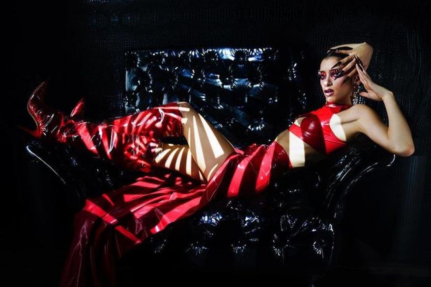 Adult female model in red vinyl dress and thigh-high boots poses on a black tufted chair, lit by light and shadows in studio.