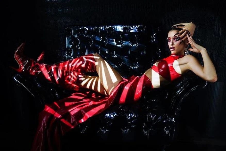 Adult female model in red vinyl dress and thigh-high boots poses on a black tufted chair, lit by light and shadows in studio.