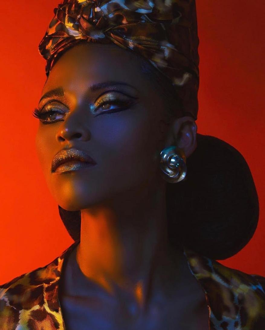 Glamorous portrait of a Black woman with dramatic makeup and an animal-print turban in a studio setting with warm lighting.