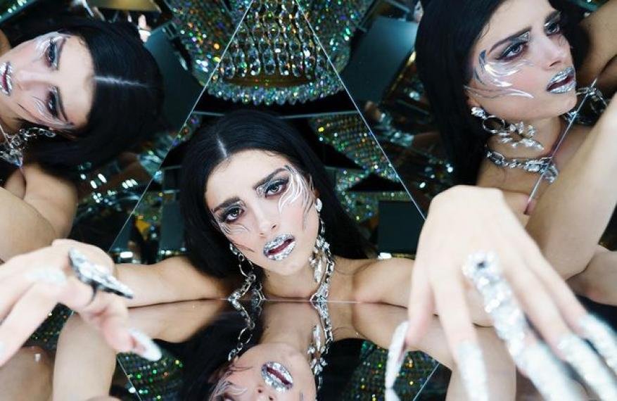 Woman in silver face makeup with long nails and jewelry strikes pose in mirrored setting with chandelier backdrop, high-fashion beauty shot.