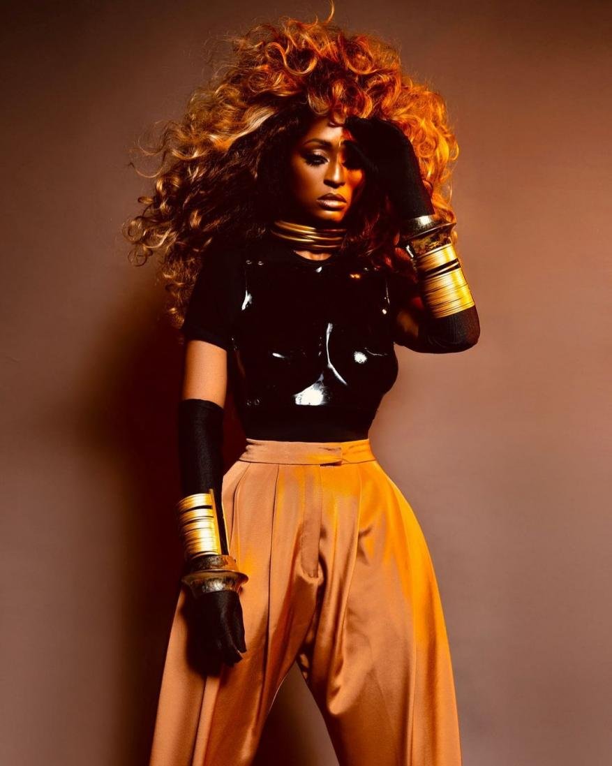 A woman with curly brown hair, wearing gold bracelets and a black top, stands against a neutral background in a fashion style photo.