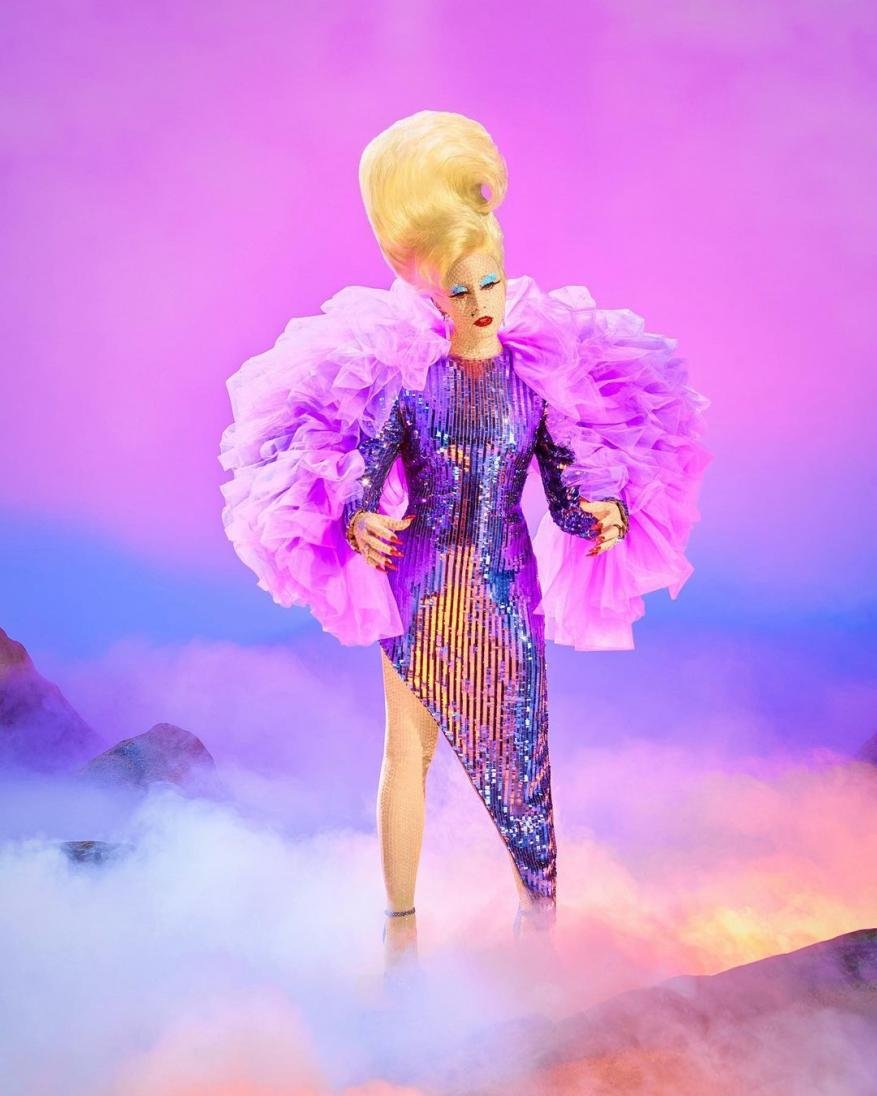 A person in a sequined gown with a large blonde beehive hairstyle and pink ruffled shoulders stands against a purple background.