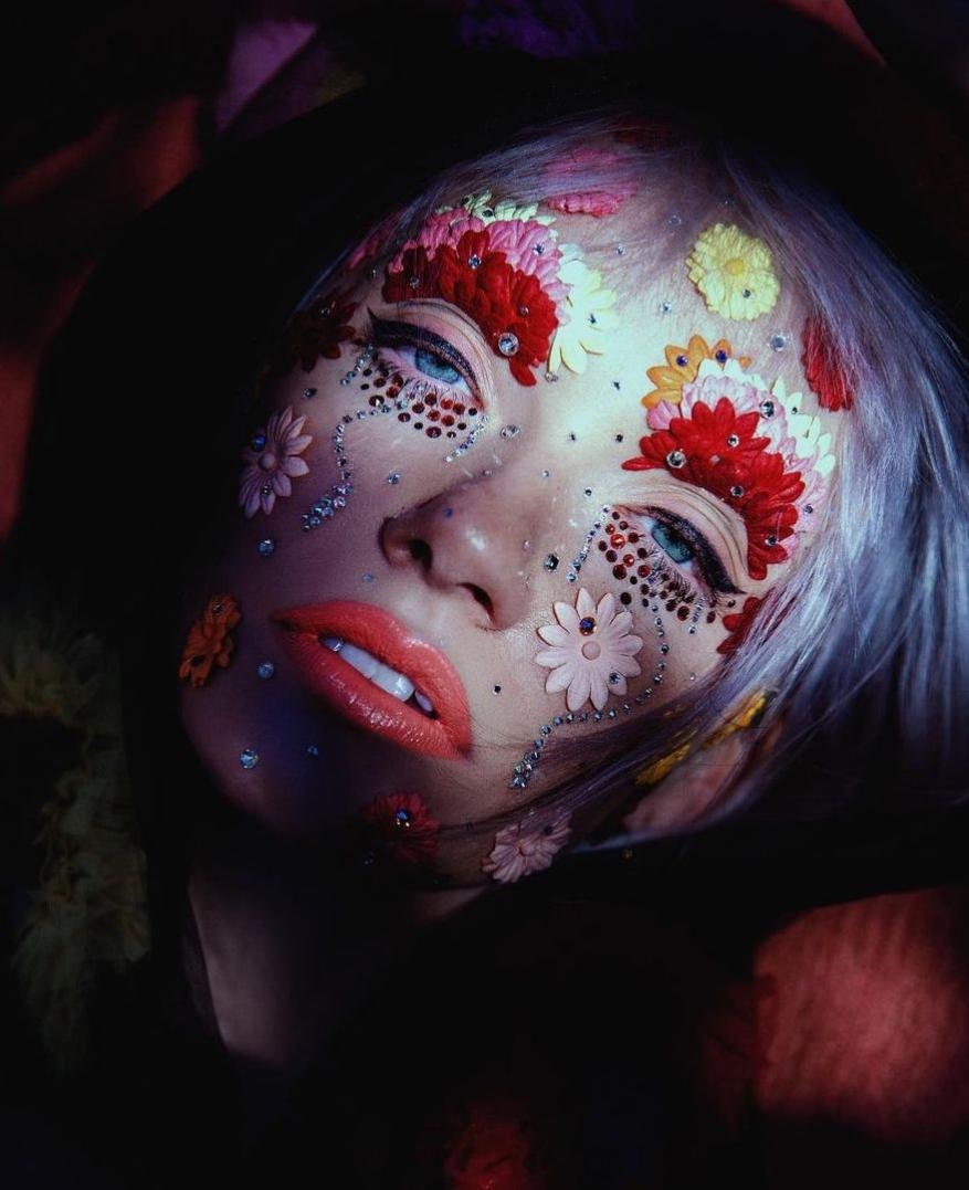Close-up beauty shot of an adult female with dramatic floral and gemstone makeup, light hair, and red lipstick against a dark floral background.