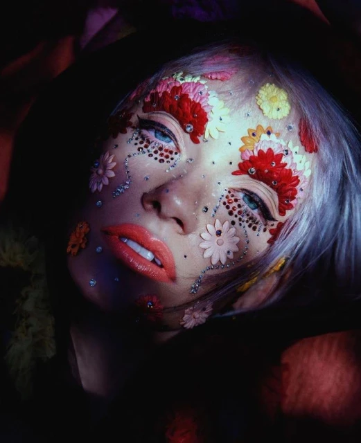 Close-up beauty shot of an adult female with dramatic floral and gemstone makeup, light hair, and red lipstick against a dark floral background.
