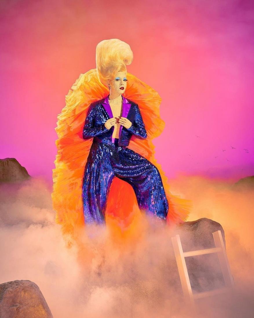 Drag queen with vibrant hair and sequin jumpsuit poses in a surreal landscape with clouds, rocks, and a ladder against a colorful pink gradient backdrop.
