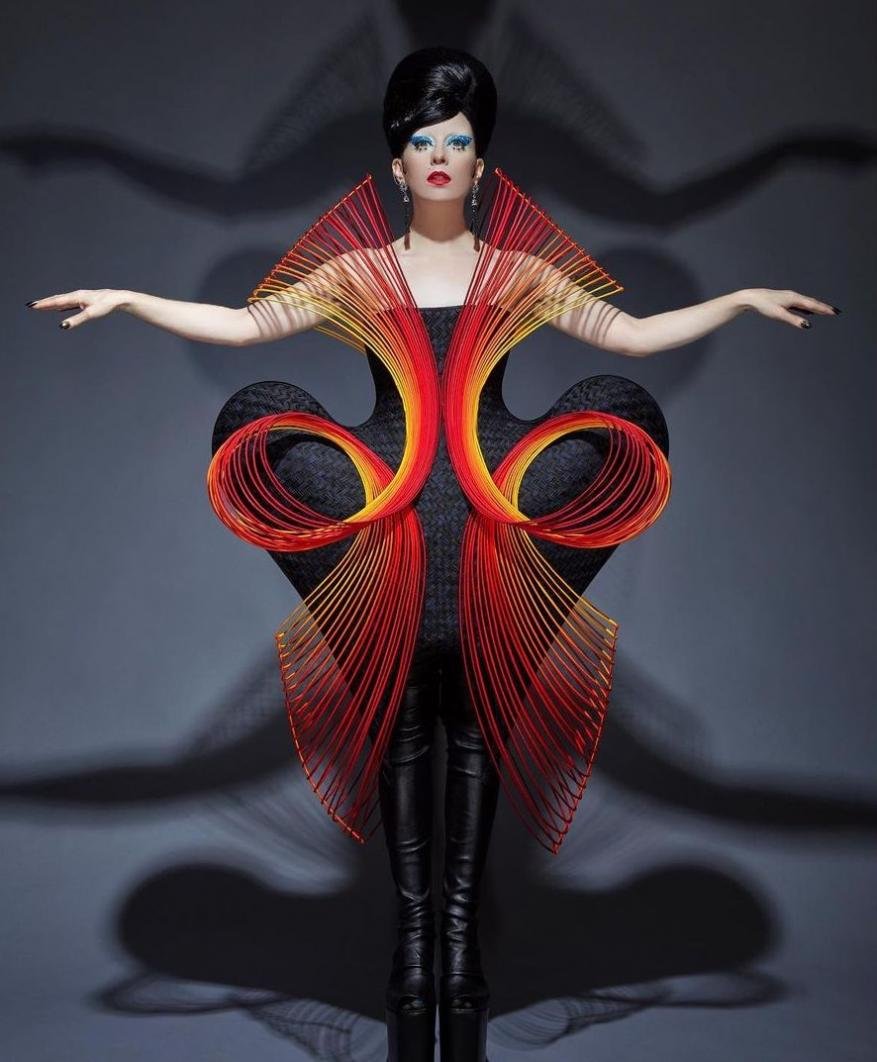 Woman posing in black dress with red and yellow string art, wearing black boots and dramatic makeup against a grey background.