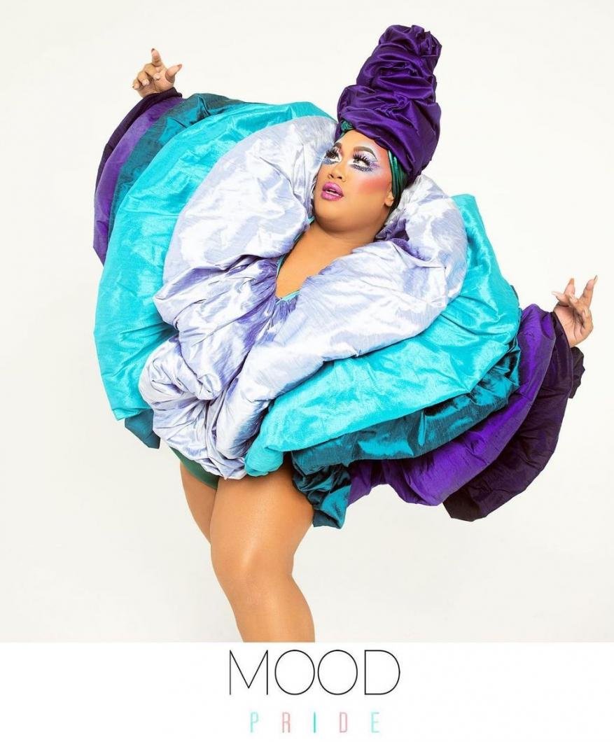Adult wearing a bright turquoise, lavender and purple puffy gown with a large head wrap in front of a white background in a studio.