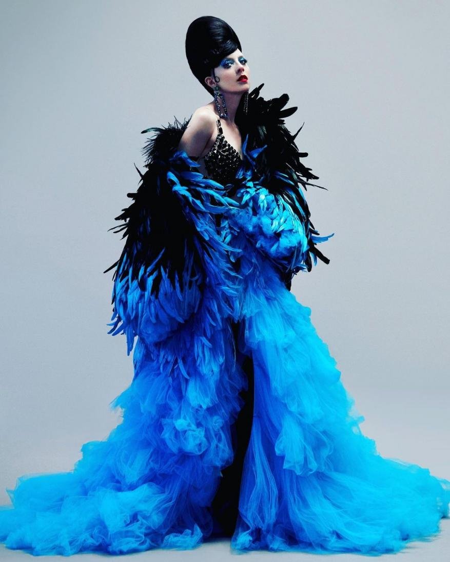 Glamorous woman with dark hair, striking makeup, and feather boa posing in a photo studio with a soft grey backdrop.