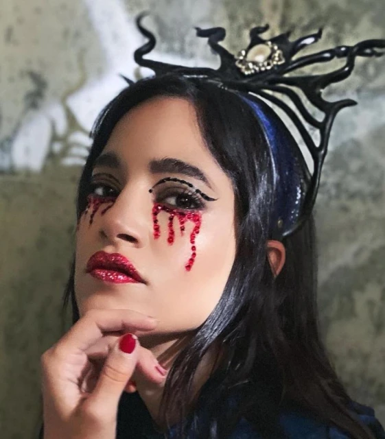Close-up of a woman with black hair wearing a jeweled crown and dramatic red makeup, her hand gently touching her chin.