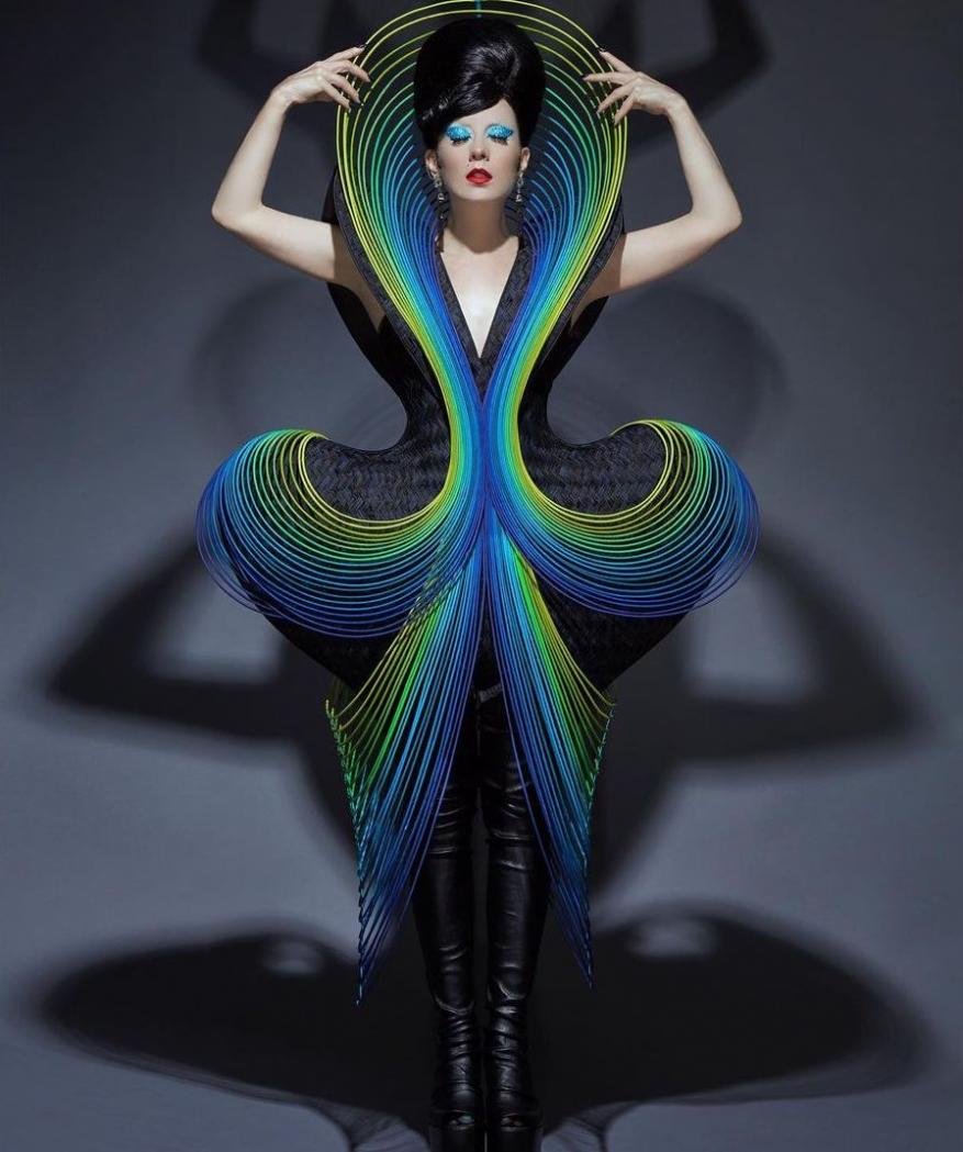 Woman in futuristic dress with blue and green curved elements, black leather pants and high heels, posing in a grey studio.