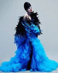 Model in voluminous blue tulle gown with feathered black shrug and tall beehive updo against a grey studio backdrop.