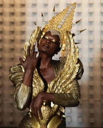 Model in ornate gold metallic headpiece and textured golden dress with dramatic winged shoulders posing against patterned wall