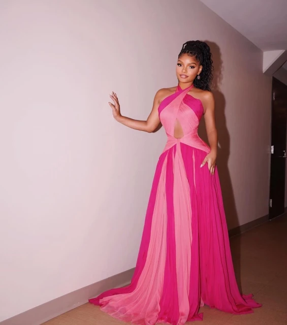 A solo shot of an adult female posing in an elegant pink gown with a train, against a plain wall, with soft indoor lighting.
