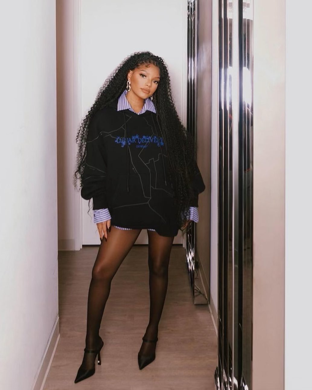 Woman poses indoors wearing a black sweatshirt dress, striped collared shirt, black tights, and black high heels near a silver accent wall.