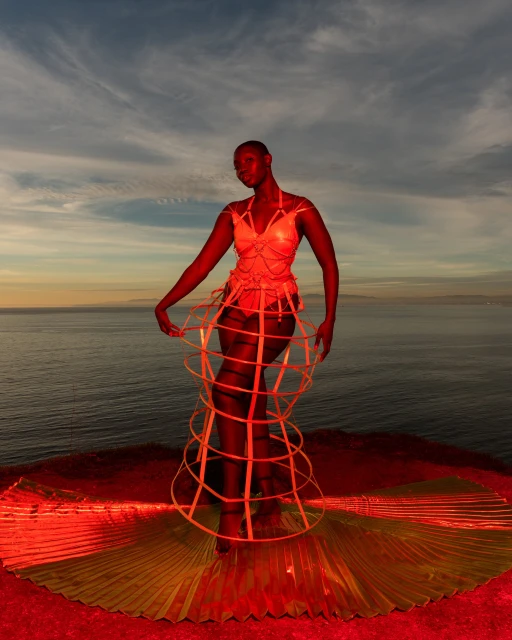 Woman poses in a sculptural cage dress on a cliff overlooking the ocean at sunset in a vibrant fashion editorial, showcasing artistic expression.