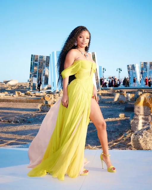 Halle Bailey poses in a yellow off-shoulder gown with a thigh-high slit, accessorized with jewelry and matching yellow heels, on a stone surface.