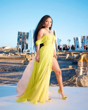 Halle Bailey poses in a yellow off-shoulder gown with a thigh-high slit, accessorized with jewelry and matching yellow heels, on a stone surface.