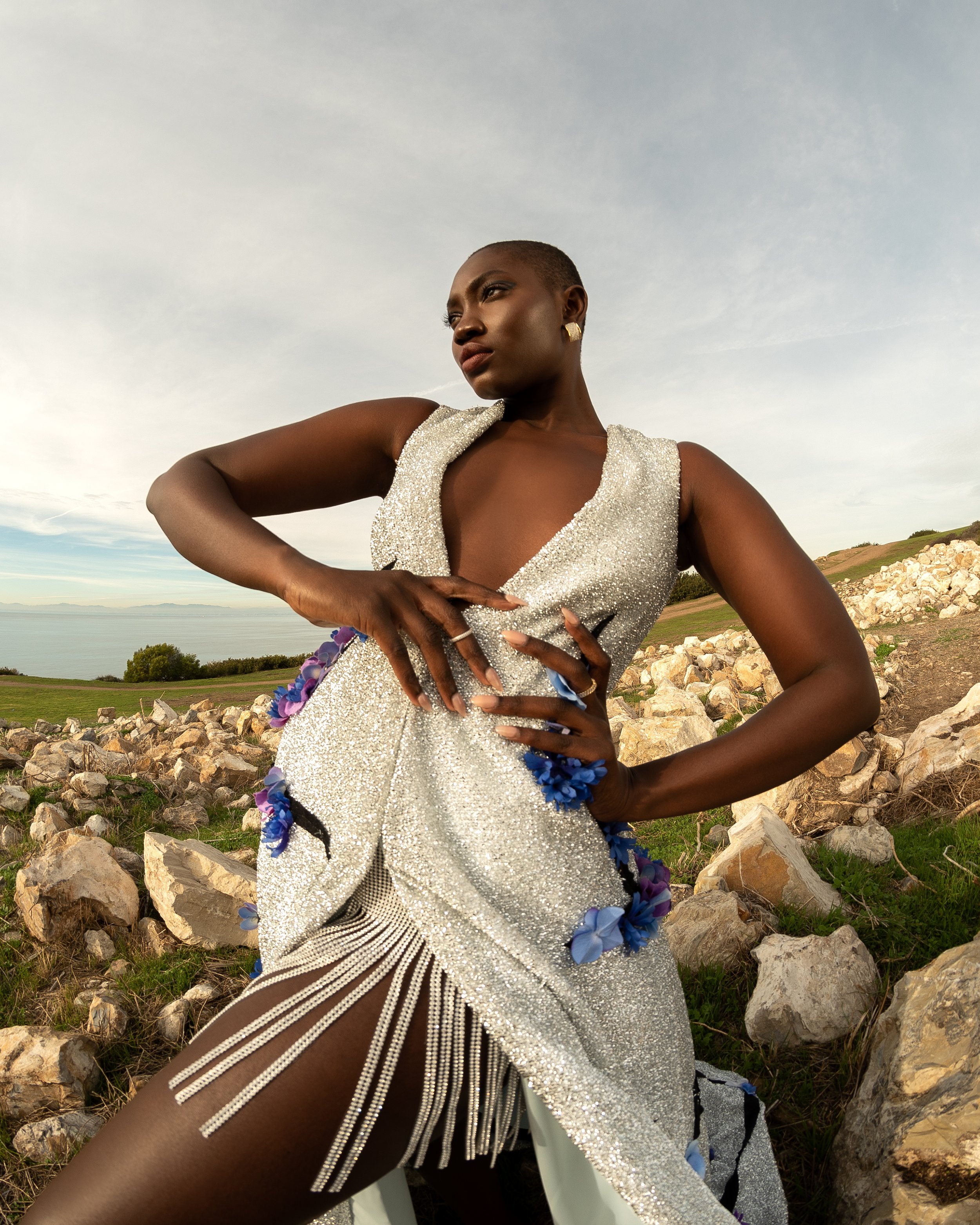 Elegant African woman in a silver, floral-accented gown poses outdoors against a natural, rocky backdrop under soft, diffused sunlight.