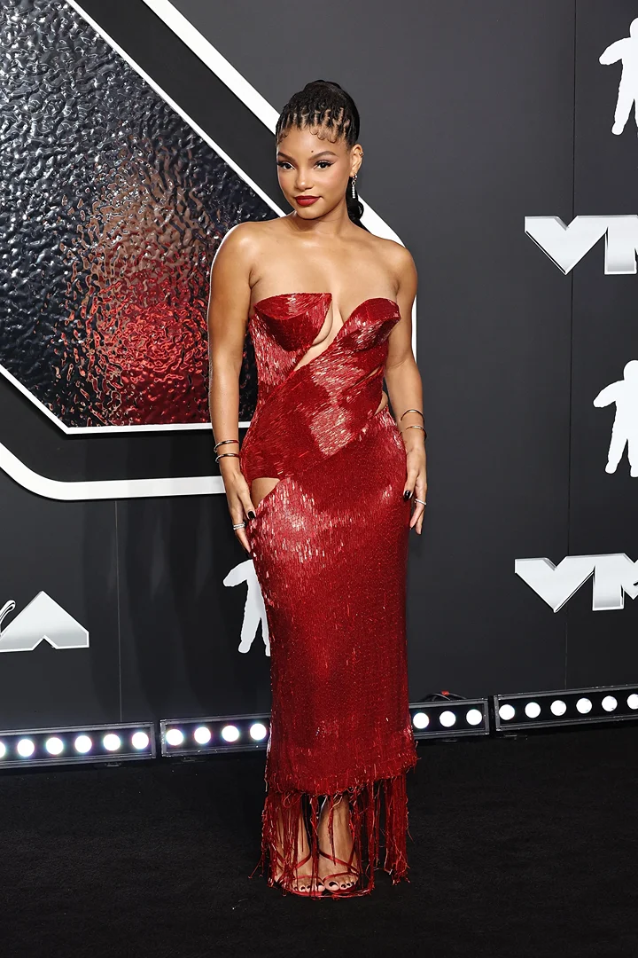 Woman in a strapless red dress on a red carpet with a geometric black and gray background and stage lighting for an event.