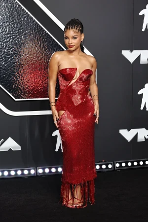 Woman in a strapless red dress on a red carpet with a geometric black and gray background and stage lighting for an event.
