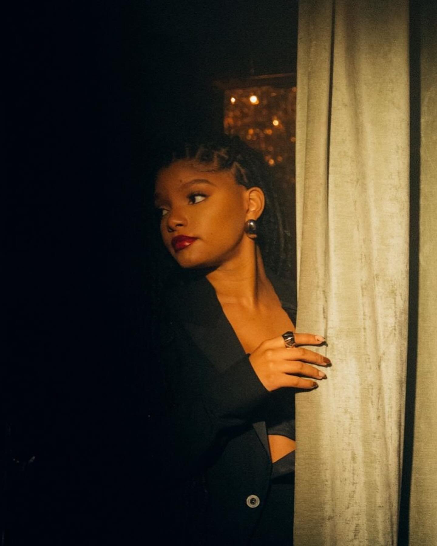 Portrait of a woman with locs, dark suit, and dramatic red lipstick, partially obscured by a gold curtain, standing indoors.