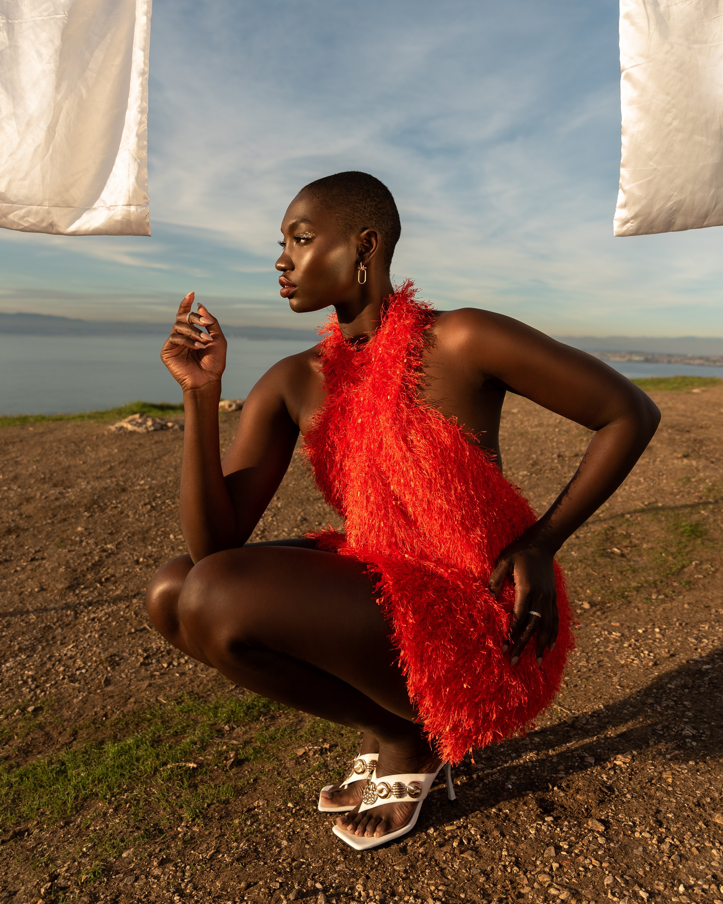 A dark-skinned woman in a bright orange dress and white heels squats on a grassy hill overlooking the ocean with a white sheet overhead.
