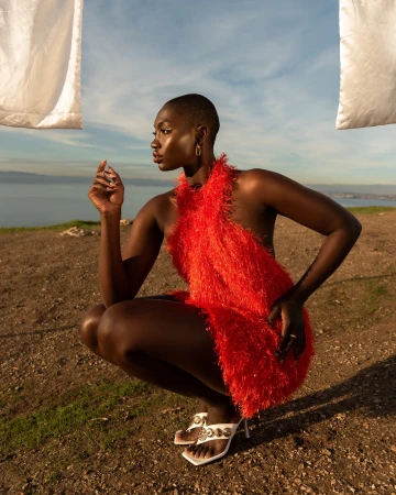 A dark-skinned woman in a bright orange dress and white heels squats on a grassy hill overlooking the ocean with a white sheet overhead.