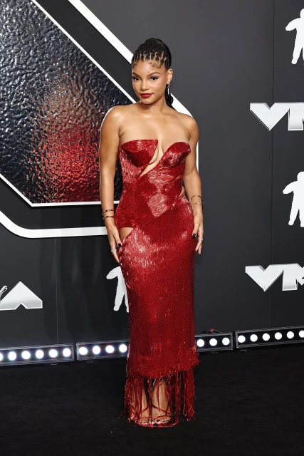 Woman on a red carpet in a floor-length strapless red gown with fringe at the bottom and dark hair, wearing jewelry.