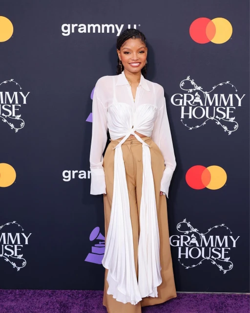Halle Bailey poses on a purple carpet in a white shirt, tan pants, and jewelry at the 2023 Grammy House event with logos in the background.