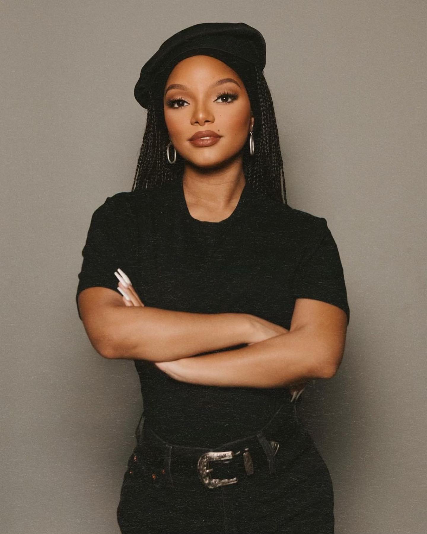 Portrait of a woman with braided hair wearing a beret, black top, and jeans, standing in front of a gray background with her arms crossed.